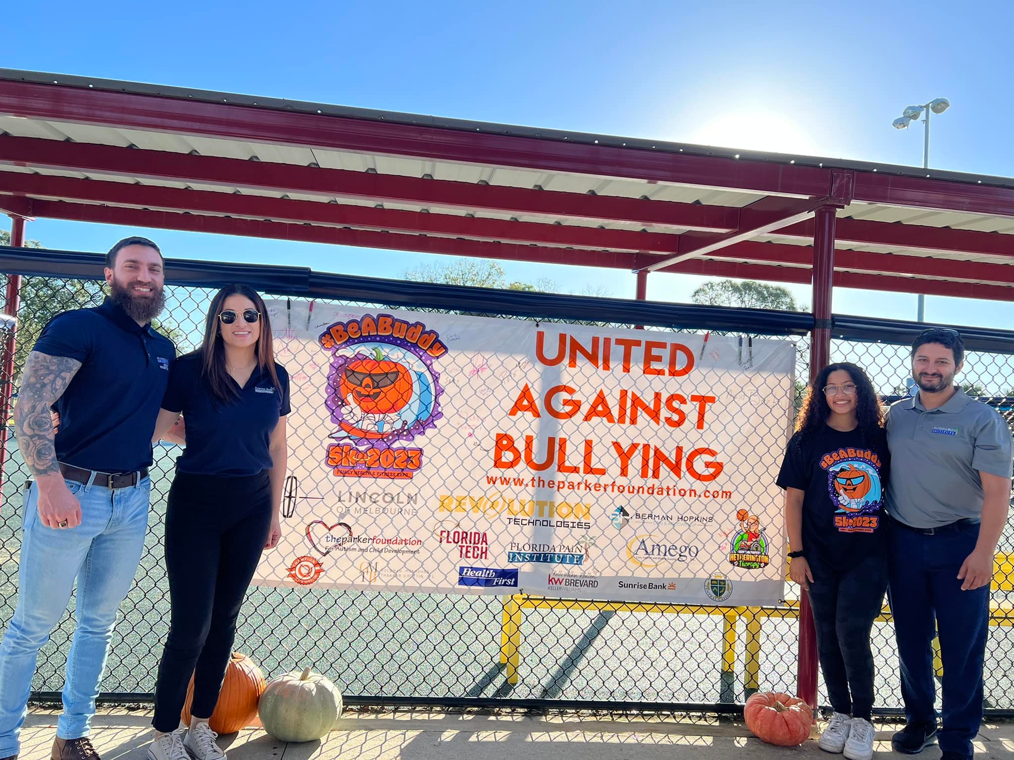 A pair of couples (one man and one woman) stand on either side of a banner hung on a chain link fence that says United Against Bullying. Three pumpkins are on the ground below.
