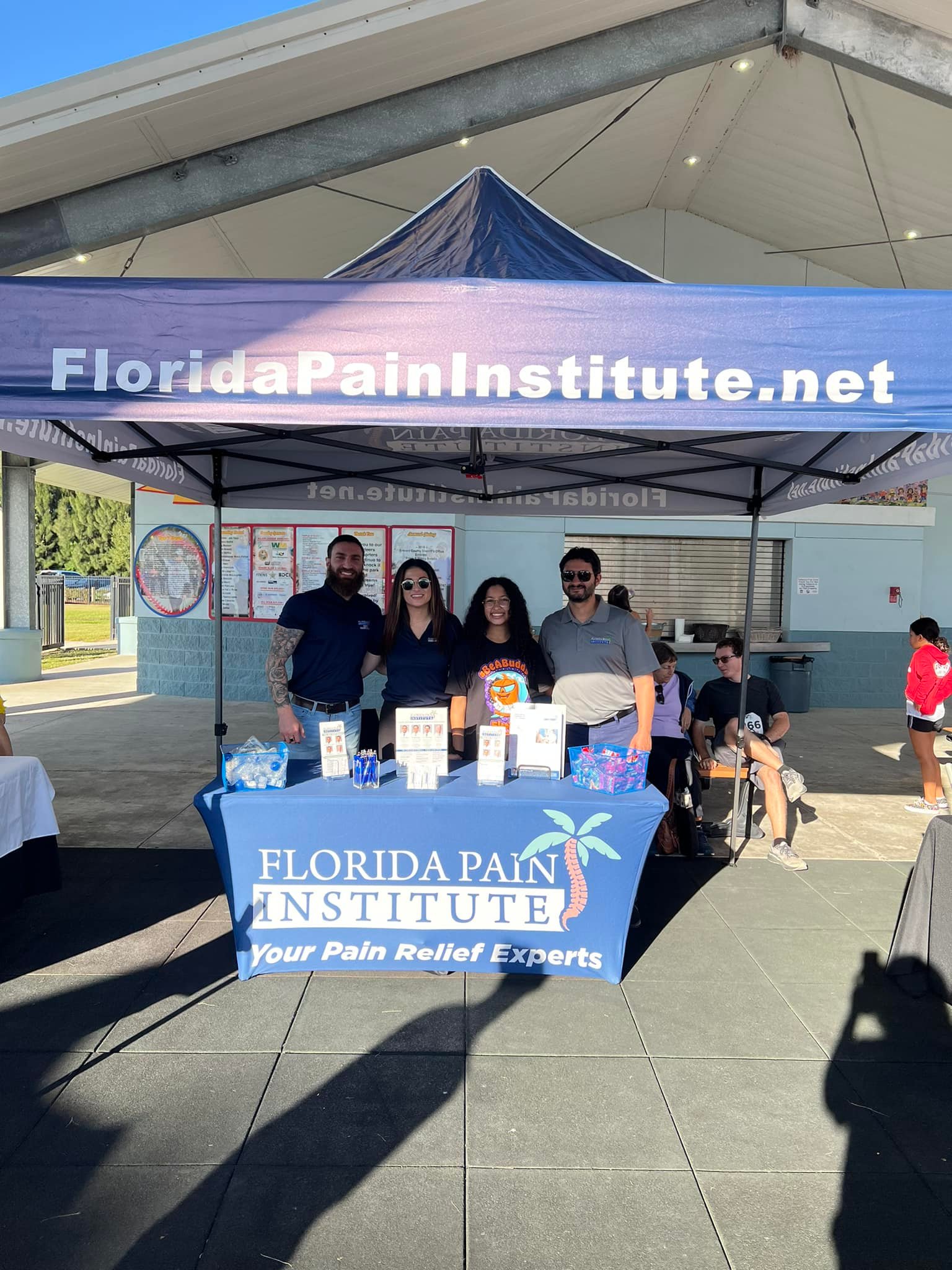 Two men and two women stand behind a table underneath a large tent. The tent and table banner are labeled Florida Pain Institute.